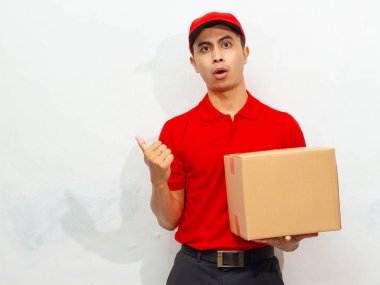 Kırmızı üniformalı ve şapkalı Asyalı erkek kurye bir paket kutusu tutuyor, baş parmağıyla gülümsüyor ve beyaz bir arka planda izole edilmiş bir şekilde kenarı işaret ediyor..