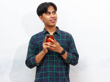 Akıllı telefon tutan yakışıklı genç bir Asyalı beyaz arka planda izole edilmiş düşünceli bir hareketle yukarı bakıyor. Düşünce, fikir ya da ilham gibi kavramlar için mükemmel..