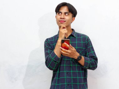 Akıllı telefon tutan yakışıklı genç bir Asyalı beyaz arka planda izole edilmiş düşünceli bir hareketle yukarı bakıyor. Düşünce, fikir ya da ilham gibi kavramlar için mükemmel..