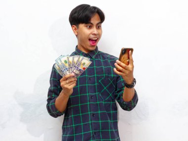 Elinde cep telefonu ve kağıt para olan yakışıklı Asyalı bir adam beyaz bir arka planda izole edilmiş bir şekilde şaşırtıcı bir ifade sergiliyor..