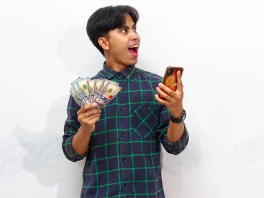 Elinde cep telefonu ve kağıt para olan yakışıklı Asyalı bir adam beyaz bir arka planda izole edilmiş bir şekilde şaşırtıcı bir ifade sergiliyor..