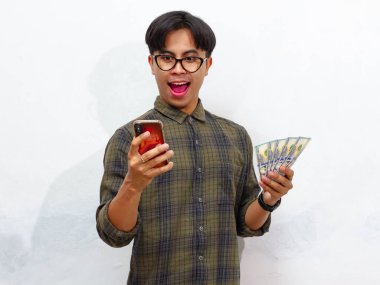 Elinde cep telefonu ve kağıt para olan yakışıklı Asyalı bir adam beyaz bir arka planda izole edilmiş bir şekilde şaşırtıcı bir ifade sergiliyor..
