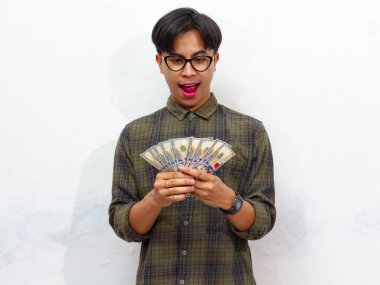 Yakışıklı Asyalı genç adam heyecanlı bir şekilde paraya bakıyor, beyaz bir arka planda izole edilmiş..