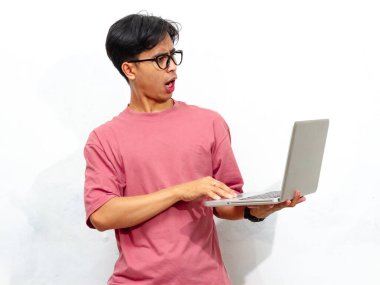 Pembe tişörtlü genç Asyalı bir erkek öğrenci. Komik bir ifadeyle beyaz arka planda dizüstü bilgisayarla poz veriyor..