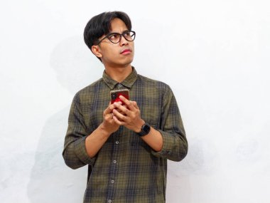 Yakışıklı Asyalı genç adam cep telefonuyla yazarken bir şey düşünüyor, beyaz bir arka planda izole edilmiş..