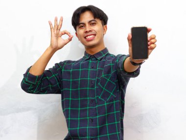 Beyaz bir arka planda izole edilmiş, mutlu, Asyalı genç bir adamın portresi elinde boş bir ekranla baş parmağını kaldırıyor ve iyi bir jest yapıyor..