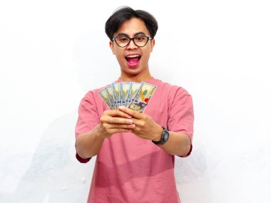 Pembe tişört giyen genç Asyalı adam elinde para tutarak poz veriyor beyaz arka planda izole edilmiş hayret verici bir ifadeyle. Sürpriz, finansal başarı ve zenginlik kavramları için mükemmel..