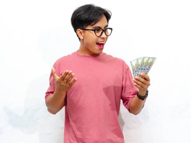 Pembe tişört giyen genç Asyalı adam elinde para tutarak poz veriyor beyaz arka planda izole edilmiş hayret verici bir ifadeyle. Sürpriz, finansal başarı ve zenginlik kavramları için mükemmel..