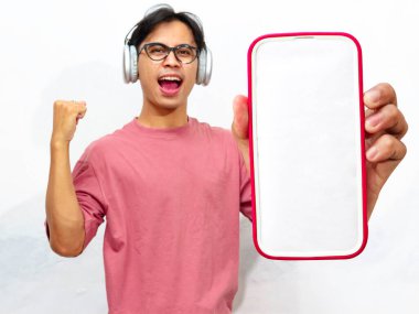 Heyecanlı genç Asyalı adam pembe tişört giymiş yumruğunu sıkıyor ve boş bir cep telefonu ekranı gösteriyor kazanan bir ifadeyle, reklam için mükemmel, beyaz arka planda izole edilmiş..