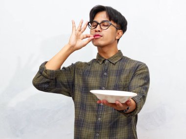 Yakışıklı Asyalı genç adam boş bir tabak tutarken gözleri kapalı lezzetli bir jest yapıyor, mutlu bir yüz ifadesi sergiliyor, beyaz bir arka planda izole edilmiş..