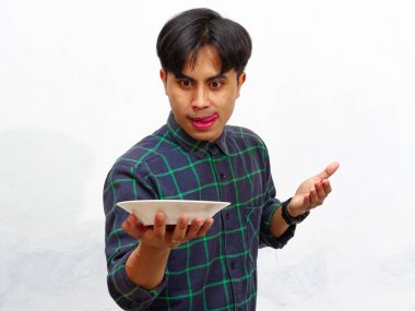 Yakışıklı bir Asyalı genç adam mutlu bir ifadeyle boş bir tabağa bakıyor, beyaz bir arka planda izole edilmiş. Yemek ve reklam kavramları için mükemmel..