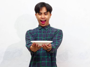 Yakışıklı bir Asyalı genç adam mutlu bir ifadeyle boş bir tabağa bakıyor, beyaz bir arka planda izole edilmiş. Yemek ve reklam kavramları için mükemmel..