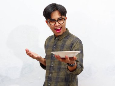 Yakışıklı bir Asyalı genç adam mutlu bir ifadeyle boş bir tabağa bakıyor, beyaz bir arka planda izole edilmiş. Yemek ve reklam kavramları için mükemmel..