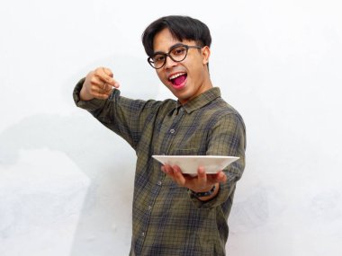 Yakışıklı Asyalı genç adam beyaz bir arka planda izole edilmiş heyecanlı ve mutlu bir ifadeyle boş bir tabağı işaret ediyor..