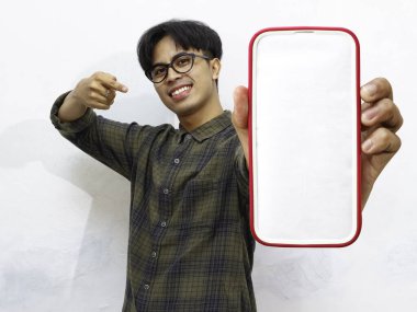 Sıradan tişörtler giyen mutlu genç Asyalı adam portresi boş ekranın akıllı telefon modelini gösteriyor ve işaret ediyor..