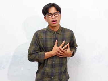 Asyalı genç bir adam sol göğsünü tutuyordu, üzgün bir ifadeyle, beyaz bir arka planda izole edilmiş..