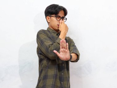 Eli ile burnunu kapatan yakışıklı Asyalı genç adam beyaz arka planda izole edilmiş bir şekilde eliyle dur işareti yapıyor..