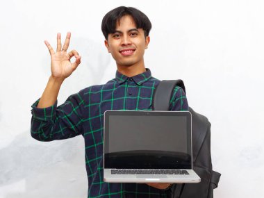 Sırt çantası tutan ve boş bir laptop ekranı gösteren kendine güvenen Asyalı erkek öğrenci, beyaz bir arka planda onay işareti ve onay veriyor..