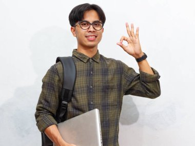 Sırt çantası takmış, elinde dizüstü bilgisayar olan, kendinden emin ve gülümseyen Asyalı bir erkek öğrenci, beyaz bir arka planda izole bir şekilde kameraya doğru işaret veriyor..