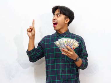 Yakışıklı Asyalı genç adam elinde para tutuyor ve beyaz arka planda izole edilmiş mutlu bir ifadeyle fikir edinme hareketini işaret ediyor.