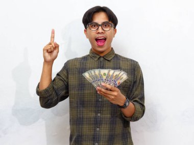 Yakışıklı Asyalı genç adam elinde para tutuyor ve beyaz arka planda izole edilmiş mutlu bir ifadeyle fikir edinme hareketini işaret ediyor.