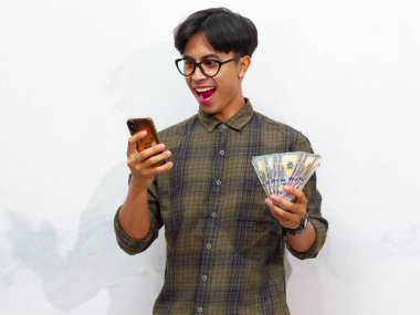 Zeki telefon ve para tutan yakışıklı genç bir Asyalı adam mutlu bir ifadeyle, beyaz bir arka planda izole edilmiş. Reklam, finansal başarı ve promosyon kullanımı için mükemmel..