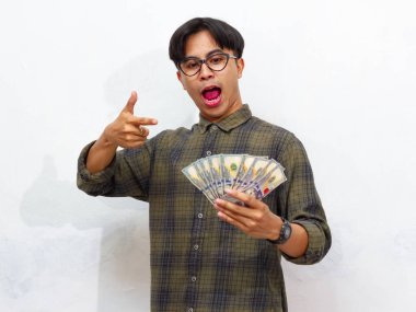 Yakışıklı Asyalı genç adam elinde kağıt para tutarken ve onu işaret ederken, beyaz bir arkaplanda yalıtılmış bir şekilde şaşırtıcı bir ifade sergiliyor..