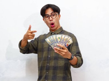 Yakışıklı Asyalı genç adam elinde kağıt para tutarken ve onu işaret ederken, beyaz bir arkaplanda yalıtılmış bir şekilde şaşırtıcı bir ifade sergiliyor..