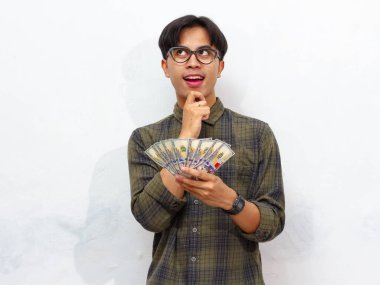 Elinde kağıt para tutan yakışıklı Asyalı bir adam beyaz bir arka planda izole bir şekilde bakan düşünceli bir jest yapıyor..