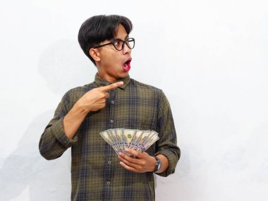 Yakışıklı Asyalı genç adam elinde kağıt para tutarken ve onu işaret ederken, beyaz bir arkaplanda yalıtılmış bir şekilde şaşırtıcı bir ifade sergiliyor..