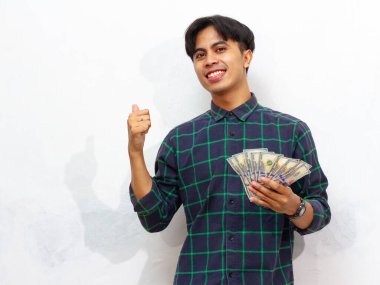 Yakışıklı Asyalı genç adam para tutuyor ve beyaz arka planda izole edilmiş gülen yüz ifadesiyle başparmaklarını kaldırıyor..