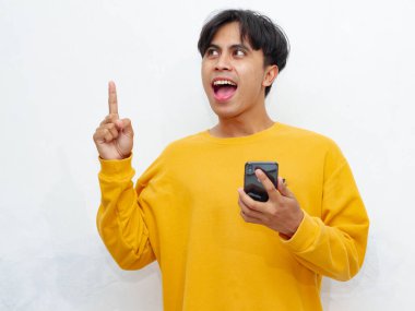 Heyecanlı Asyalı adam beyaz arka planda izole edilmiş bir cep telefonunu tutarken aklına harika bir fikir geliyor. Reklam ve yaratıcılık kavramları için ideal..