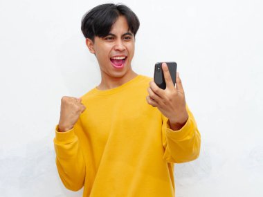 Akıllı telefonu tutan ve ona bakan heyecanlı genç adam zafer jesti yapıyor, pozitifliğini dile getiriyor ve kutluyor.