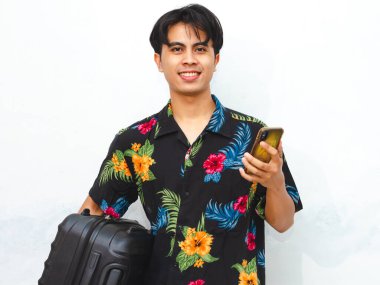 Portrait of a young Asian man wearing summer clothes, holding a mobile phone and suitcase, ready for travel abroad. Perfect for tourism, vacation, or air flight journey concepts. clipart