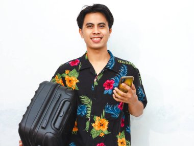 Portrait of a young Asian man wearing summer clothes, holding a mobile phone and suitcase, ready for travel abroad. Perfect for tourism, vacation, or air flight journey concepts. clipart