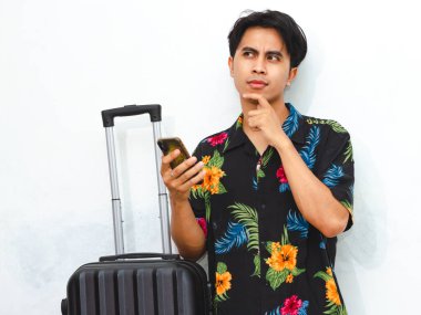 Portrait of a happy young Asian man wearing casual summer clothes, holding a mobile phone and suitcase, looking aside thoughtfully, representing a traveler ready for an adventure. clipart