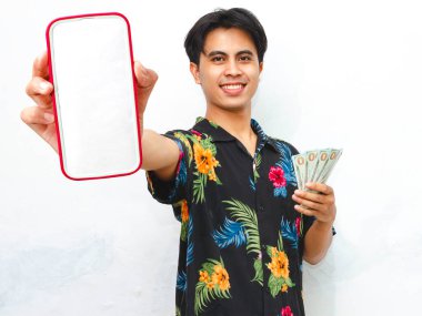 A happy young Asian man in summer casual attire shows a blank mobile phone screen while holding cash, smiling against a clean white background. clipart