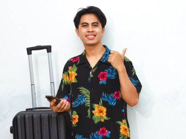 Portrait of a happy young Asian man wearing summer casual clothes, holding a phone, carrying a suitcase, and pointing with his thumb. Tourist ready for vacation. Isolated on white background. clipart