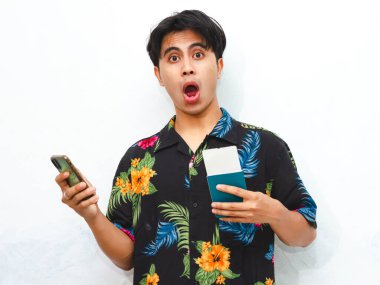 Pasaport taşıyan ve beyaz bir arka planda izole edilmiş cep telefonu kullanan sıradan bir Asyalı komik bir genç. Seyahat, tatil ve sürpriz kavramlar için mükemmel..