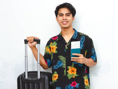 Pasaport ve bavul taşıyan yaz kıyafeti giymiş neşeli Asyalı genç bir adam yolculuğa hazır. Beyaz bir arkaplanda izole edilmiş, seyahat temalı içerik için mükemmel.
