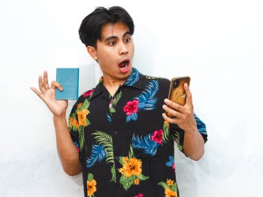 Pasaport taşıyan ve beyaz bir arka planda izole edilmiş cep telefonu kullanan sıradan bir Asyalı komik bir genç. Seyahat, tatil ve sürpriz kavramlar için mükemmel..