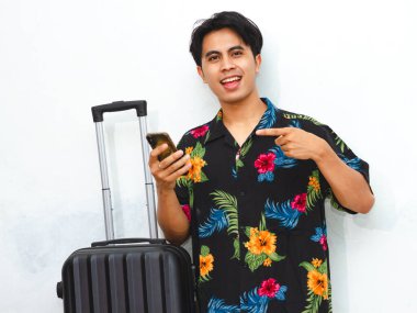 Portrait of a happy young Asian man wearing summer casual clothes, holding a phone and suitcase, pointing to copy space. Tourist traveling abroad on vacation, isolated on white. clipart