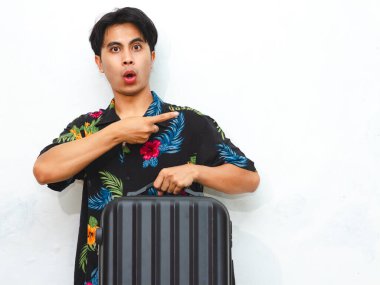 Portrait of a young Asian man in casual summer clothes carrying a suitcase, pointing aside with a surprised expression. Ideal for travel, tourism, or vacation-related ads. clipart