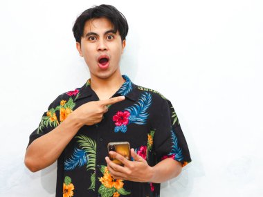 Yazın giyinmiş komik bir Asyalı genç adam beyaz arka planda izole edilmiş bir cep telefonu kullanırken yan tarafı işaret ediyor..