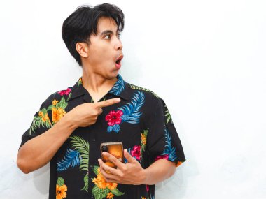 Yazın giyinmiş komik bir Asyalı genç adam beyaz arka planda izole edilmiş bir cep telefonu kullanırken yan tarafı işaret ediyor..