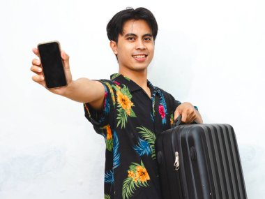 Young Asian man in summer casual wear joyfully shows a blank mobile phone screen while carrying a suitcase, embodying the spirit of travel and adventure. clipart