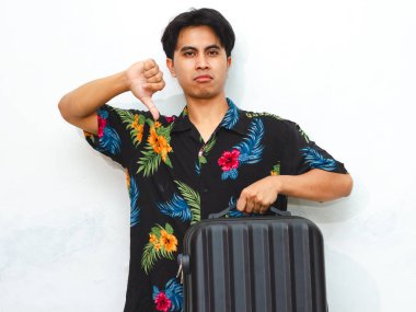 Portrait of a young Asian man in casual summer attire, holding a suitcase, giving a thumbs down with an angry expression, isolated on a white background. Tourist frustrated on vacation. clipart