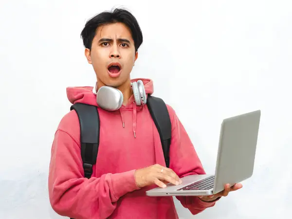 stock image Portrait of a shocked young Asian male college student wearing a pink hoodie, using a laptop, and posing on a white background.