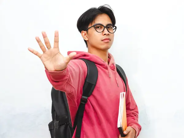 Stock image Portrait of an angry Asian male college student wearing a pink hoodie and carrying a backpack, giving a stop gesture with his hand, isolated on a white background.