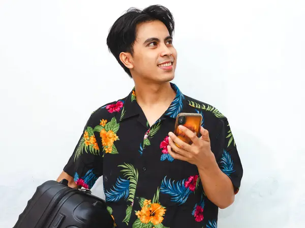 stock image Portrait of a happy young Asian man wearing casual summer clothes, holding a mobile phone and suitcase, looking aside thoughtfully, representing a traveler ready for an adventure.